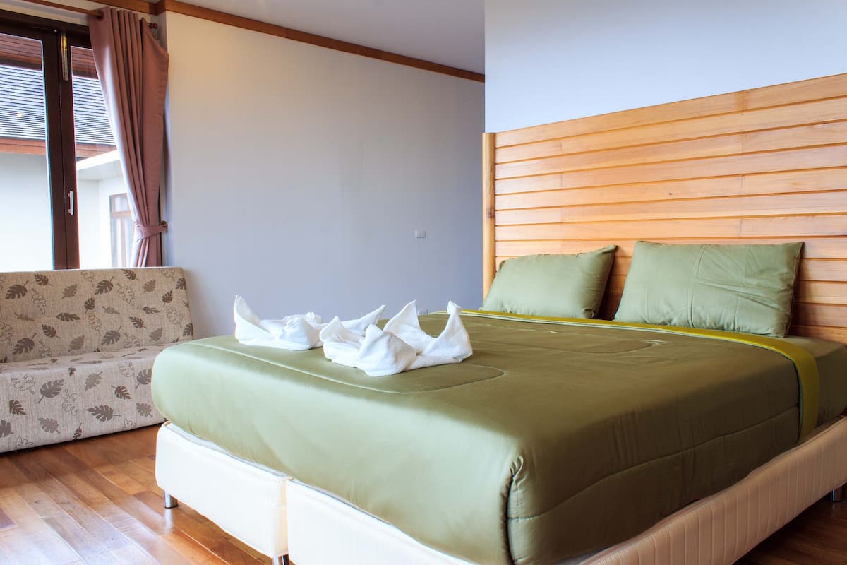 Una cama bien hecha con ropa de cama y almohadas verdes, un cabecero de madera y un sofá estampado con motivos florales en una habitación con piso de madera.