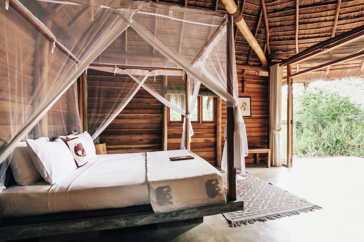 Eine Holzhütte mit einem Bett unter einem Moskitonetz, einer Holzbank und Blick auf grüne Natur.