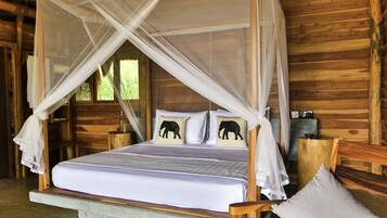 A wooden cabin bedroom with a four-poster bed, two elephant-themed pillows, and a mosquito net.