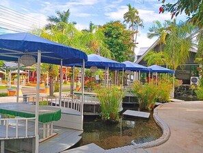 A covered outdoor seating area with tables and umbrellas, surrounded by lush greenery and a small water feature.