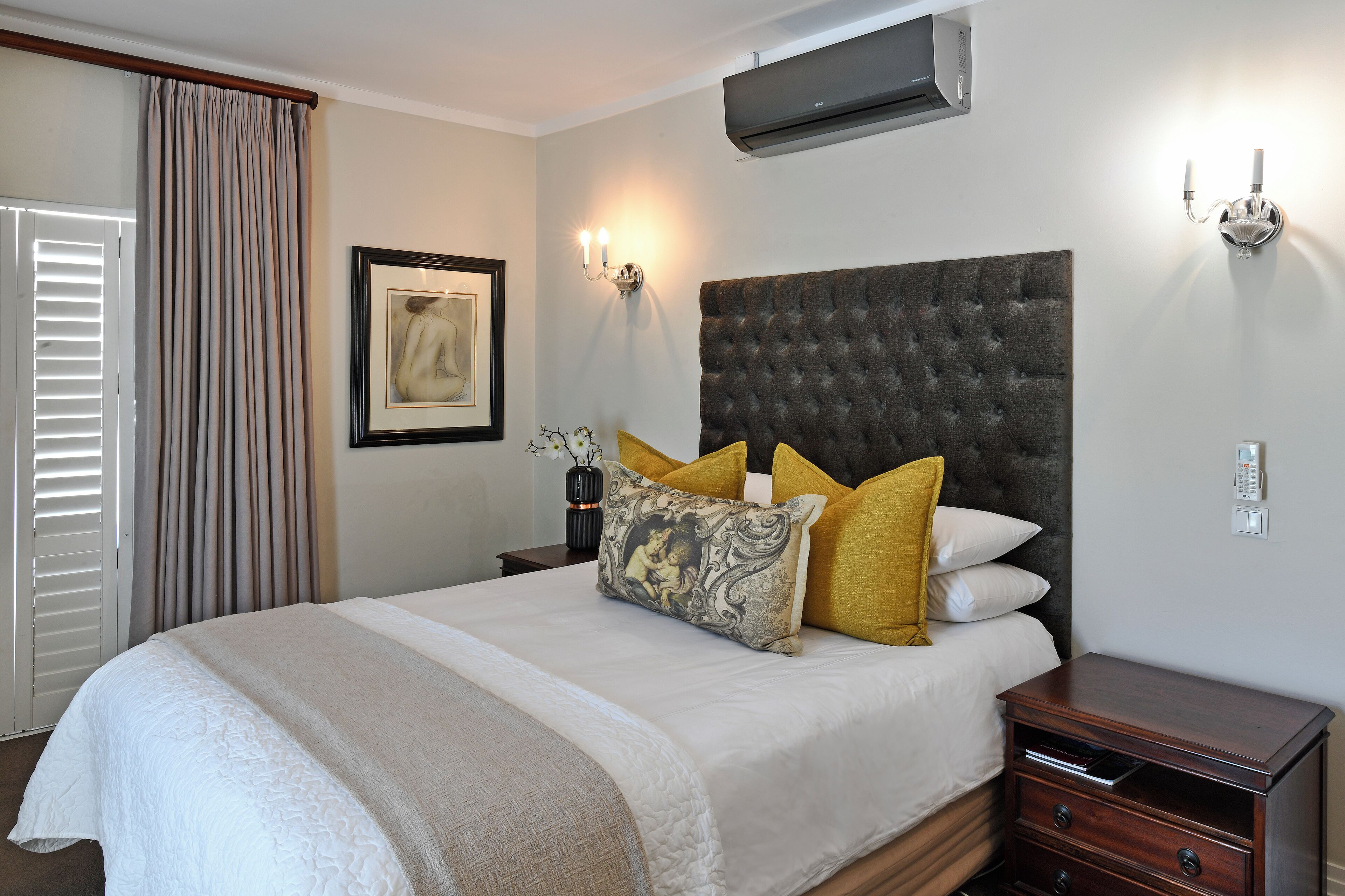 A neatly made bed with a large tufted headboard, a bedside table with a lamp, and a wall-mounted air conditioner.
