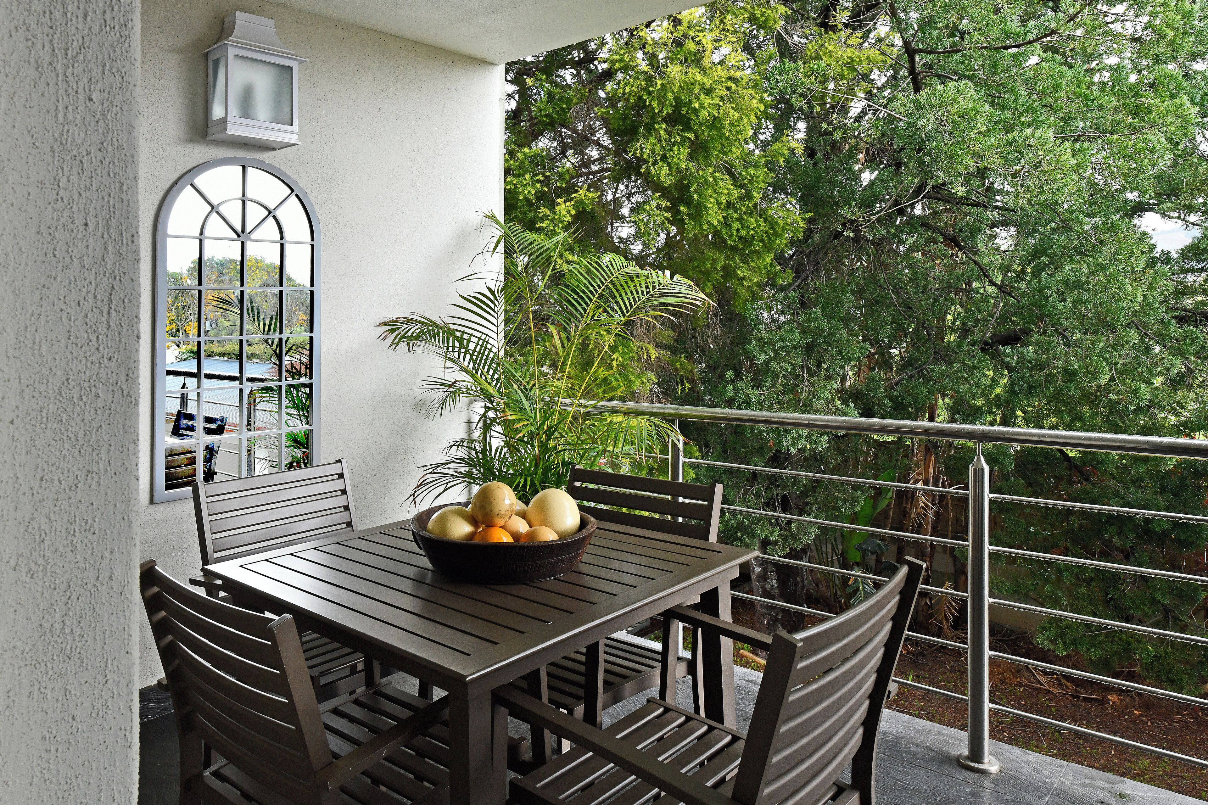 A balcony with a table and chairs, a bowl of fruit, and a view of trees.