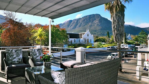 A covered outdoor seating area with wicker furniture and a view of a mountain and church. - Le Petit Bijou Boutique Aparthotel (Franschhoek)