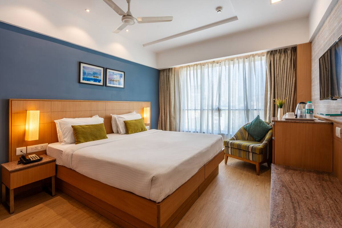 Quarto de hotel com uma cama grande, luminárias de cabeceira, uma escrivaninha com cadeira e vista para o exterior através de uma janela com cortinas.