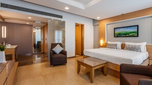 A hotel room with a large bed, a desk, a chair, and a small table. There is a painting on the wall and a door leading to another room. - GCC Northside Hotel (Mumbai)