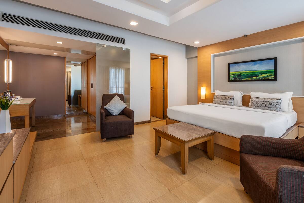 Quarto de hotel com cama grande, escrivaninha, cadeira e mesa pequena. Há um quadro na parede e uma porta que dá para outro cômodo.