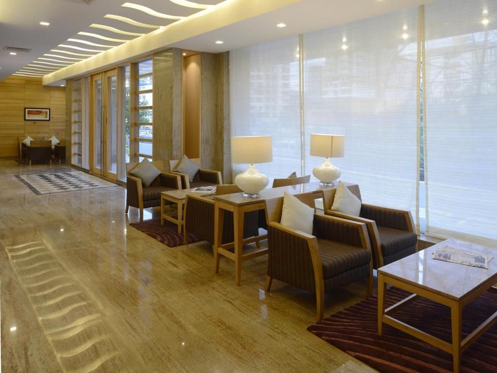 A hotel lobby with a reception desk, seating area with tables and chairs, and large windows with blinds.