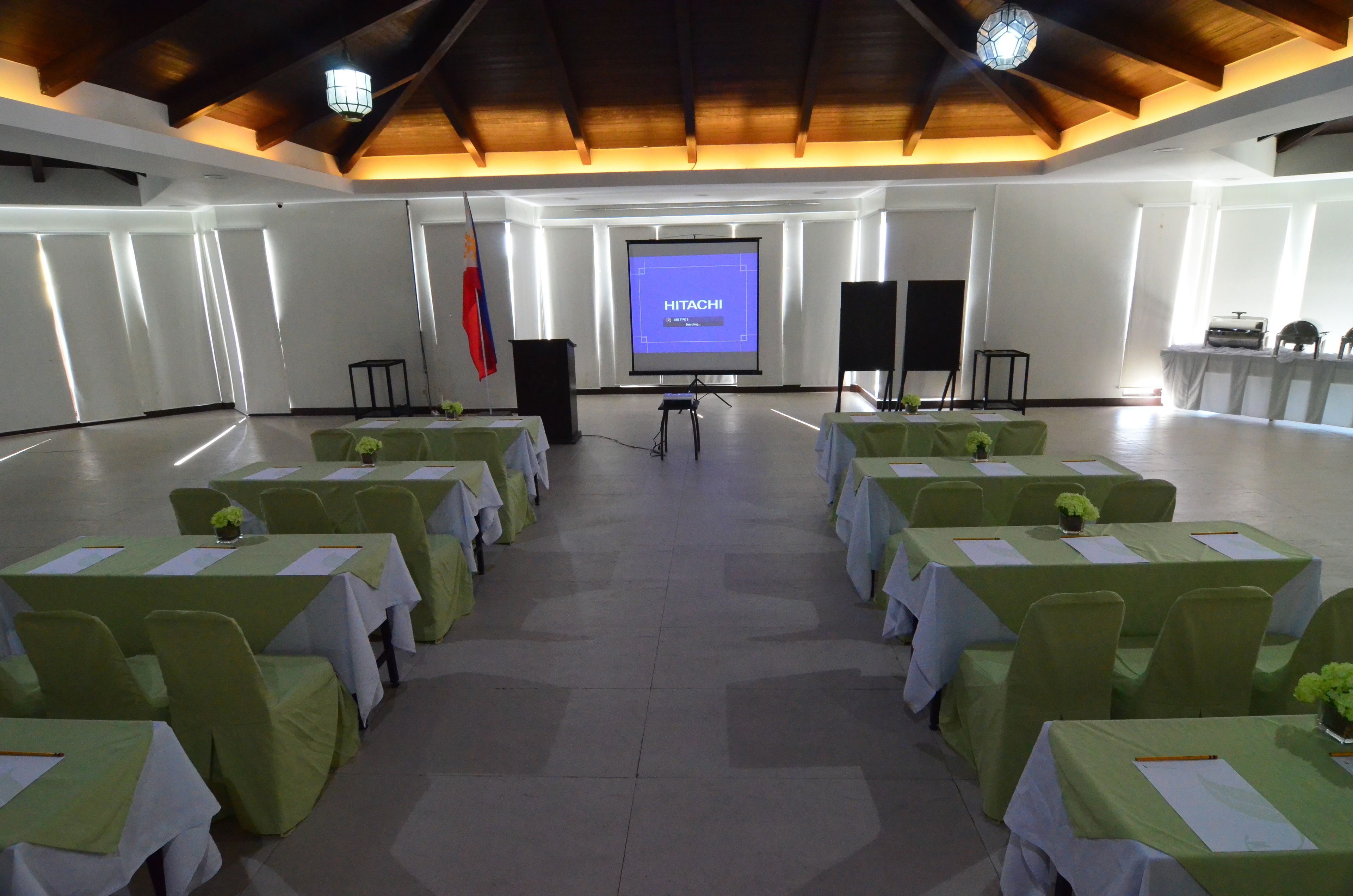 Un salón de conferencias con filas de mesas y sillas, una pantalla de proyector mostrando 