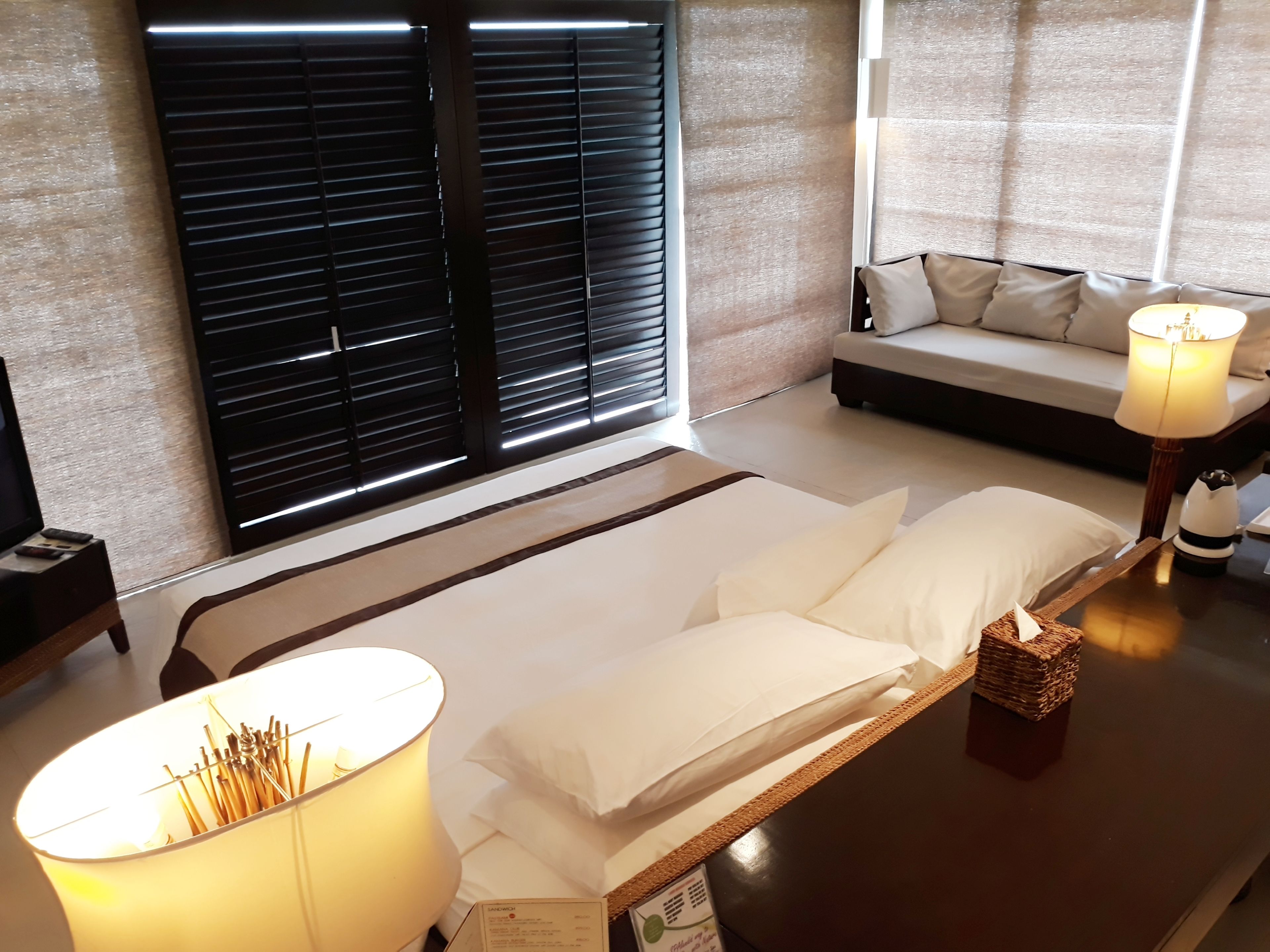 Habitación de hotel con cama, una mesita de noche con lámpara, una pequeña mesa con vela y una ventana con persianas.