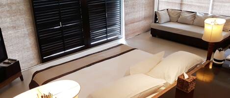 Habitación de hotel con cama, una mesita de noche con lámpara, una pequeña mesa con vela y una ventana con persianas.
