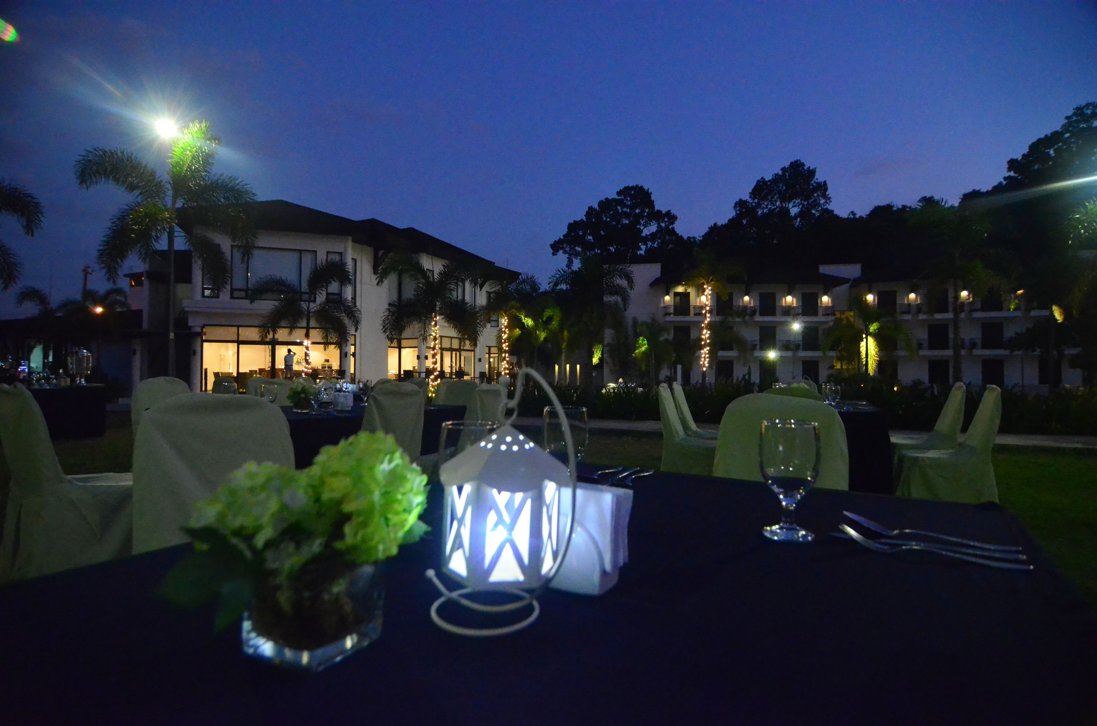 An outdoor dining area with tables set for a meal, palm trees, and a building with lights in the background.