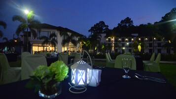 An outdoor dining area with tables set for a meal, palm trees, and a building with lights in the background.