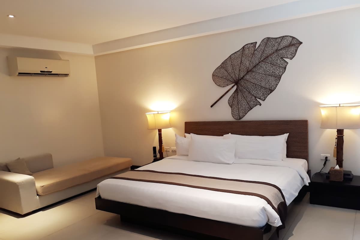Une chambre d’hôtel avec un grand lit, deux lampes de chevet, un climatiseur fixé au mur, un canapé et une œuvre d’art décorative représentant des feuilles.