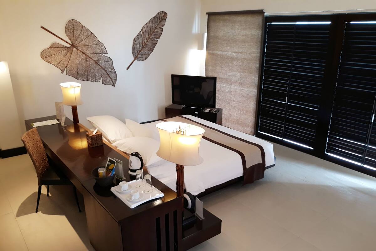Une chambre d’hôtel avec un lit, des lampes de chevet, une télévision, un bureau avec une lampe et des feuilles décoratives accrochées au mur.