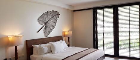 A hotel room with a large bed, two bedside lamps, a wall-mounted decorative leaf, and a large window with blinds.