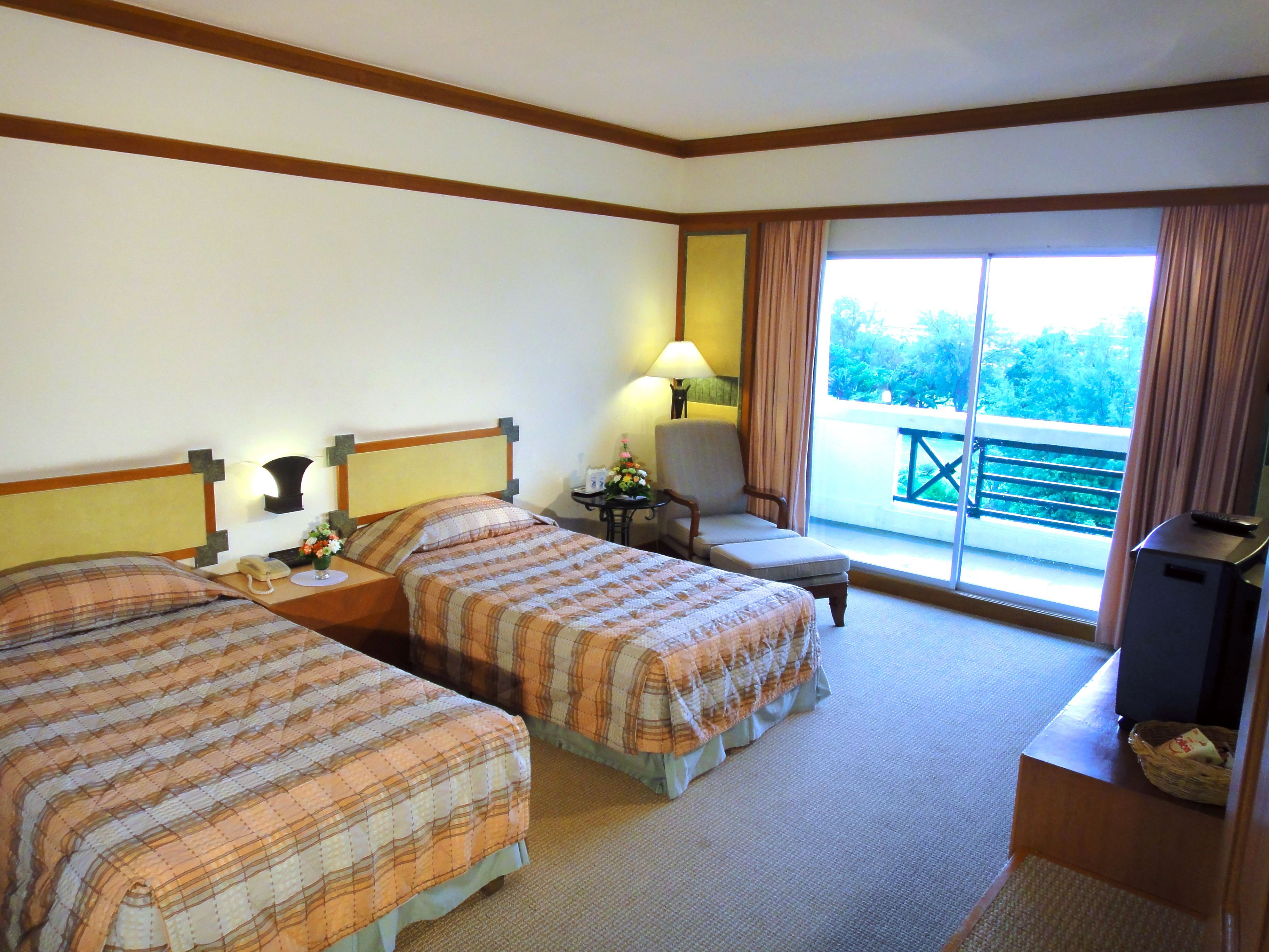Une chambre d’hôtel avec deux lits, un bureau, une chaise, une lampe et un balcon donnant sur un espace verdoyant.