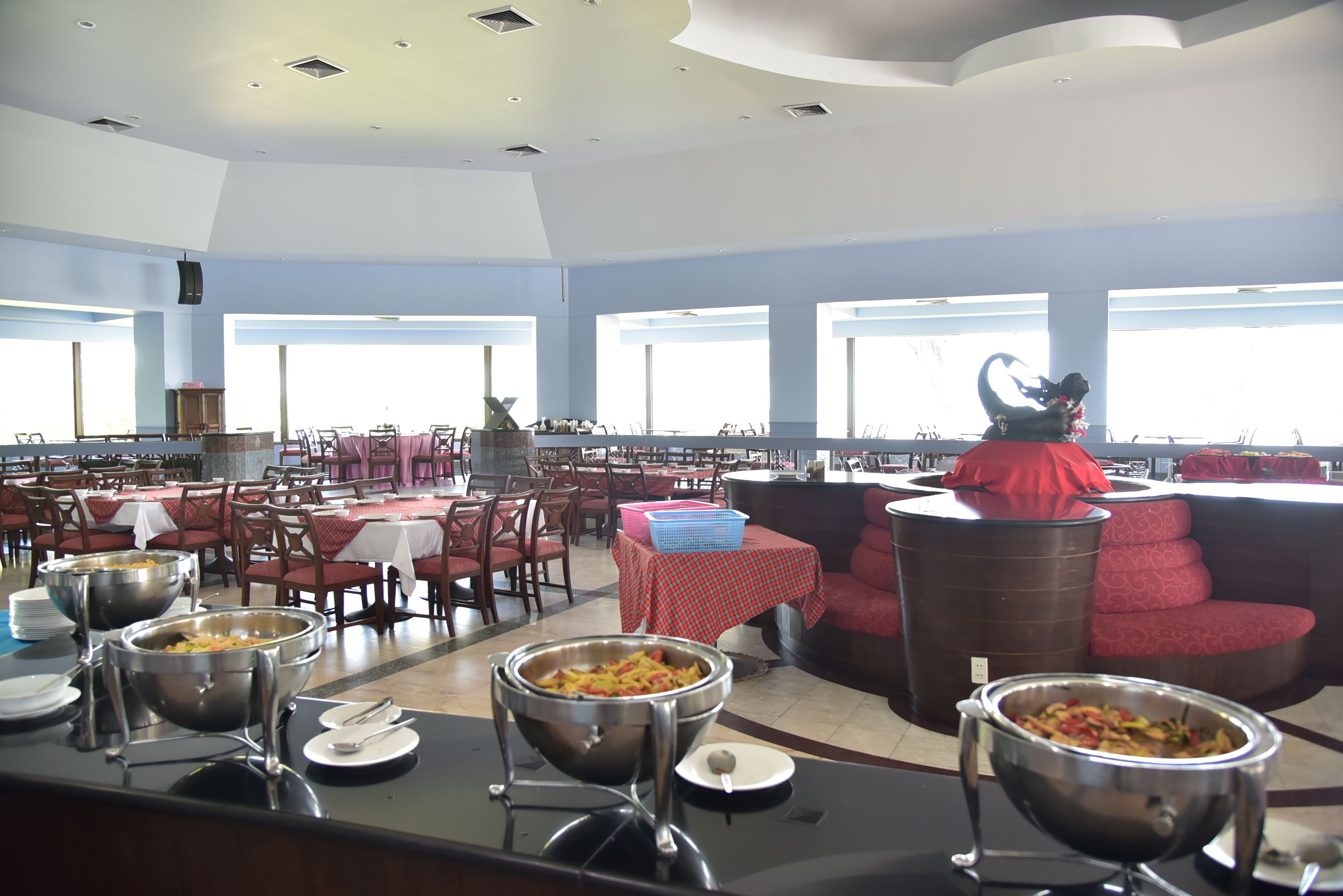 Amplia zona de comedor con múltiples mesas y sillas, estaciones de buffet con comida, y un ventanal que proporciona luz natural.