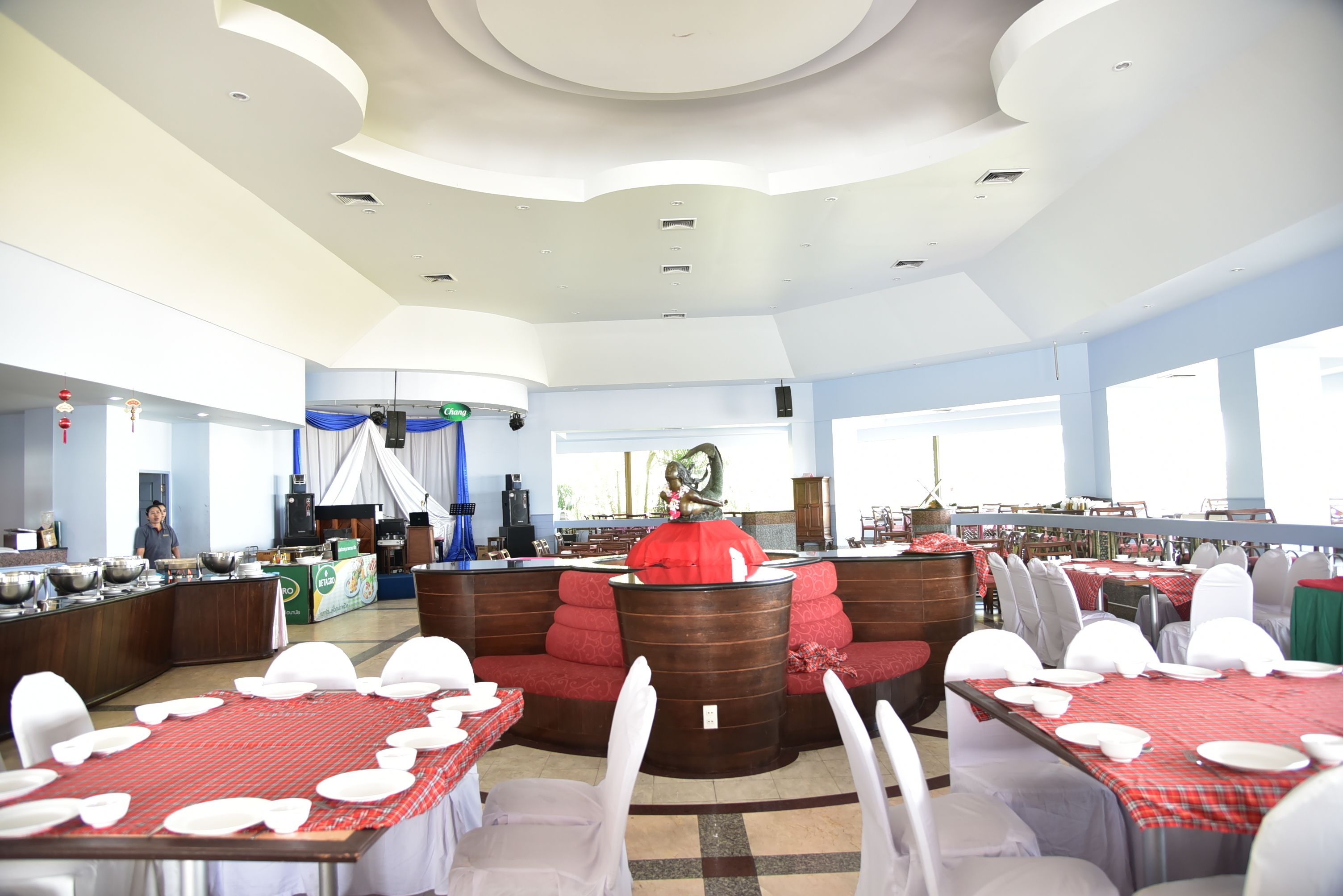 A spacious dining area with multiple tables set for guests, a central decorative feature, and a view of the outdoors.