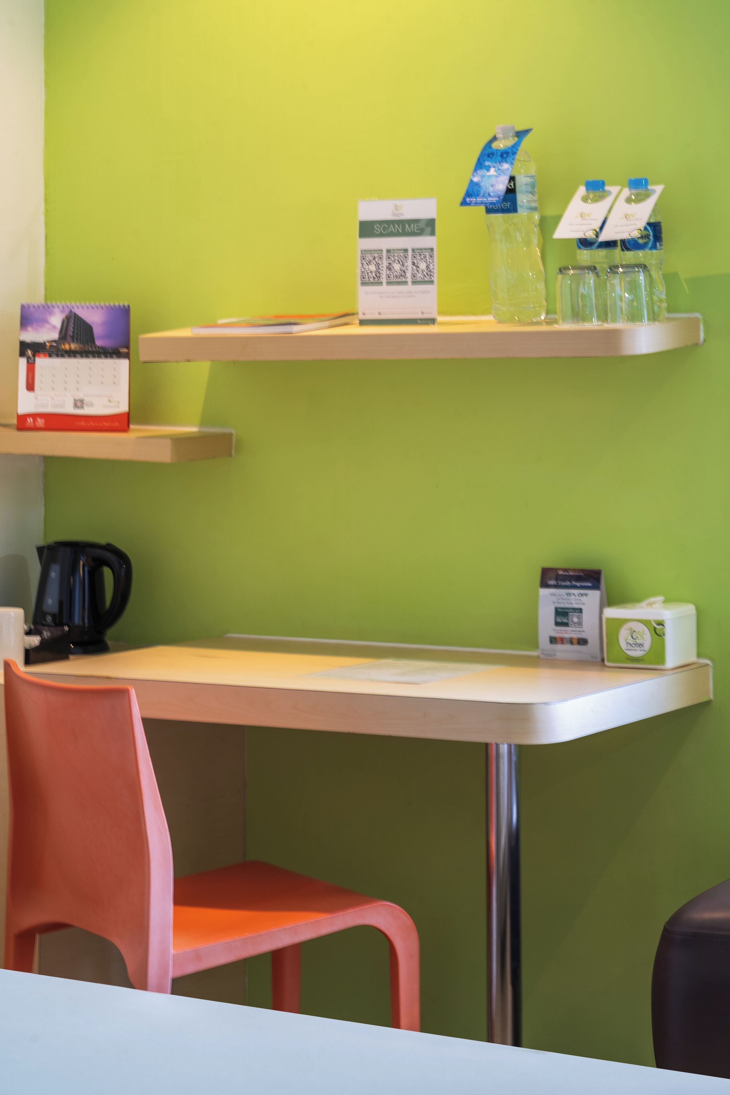 Una habitación con pared verde, una mesa blanca, una silla roja, un hervidor negro, un calendario, una tarjeta que dice 'Scan Me' y agua embotellada.