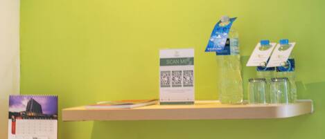 Una habitación con pared verde, una mesa blanca, una silla roja, un hervidor negro, un calendario, una tarjeta que dice 'Scan Me' y agua embotellada.