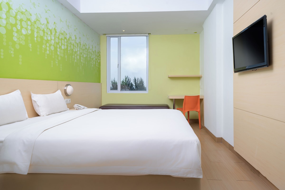 A hotel room with a large bed, a desk, a chair, a TV, and a window with a view of trees.