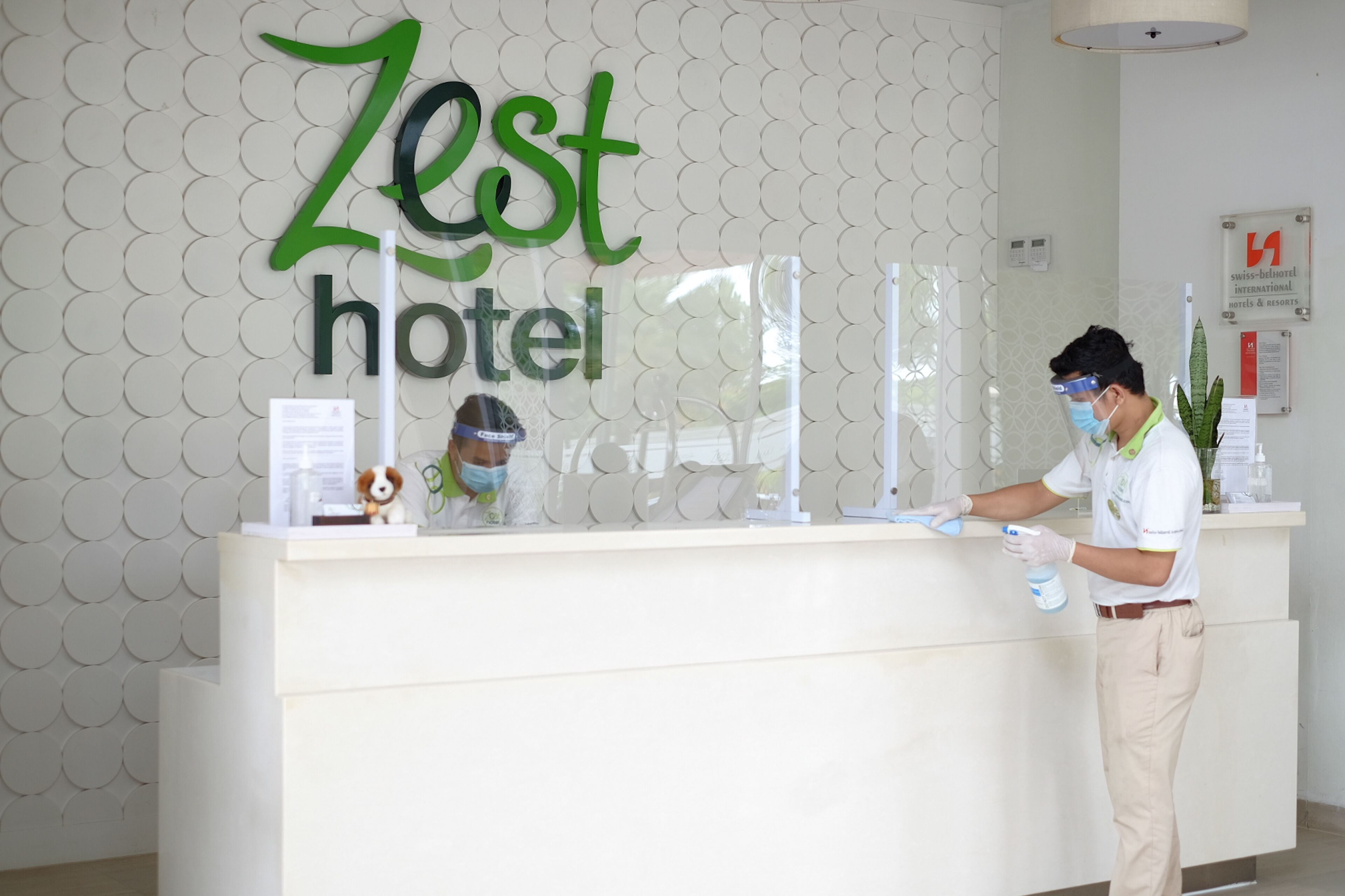 Tauleller de recepció a l'Hotel Zest amb disseny modern, un cartell visible i un membre del personal portant mascareta i guants.