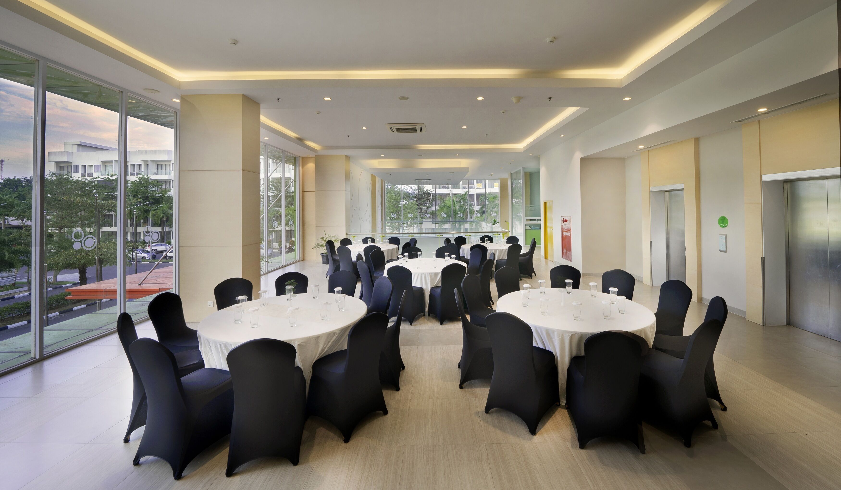 A conference room with round tables and black chairs, large windows with a city view, and a modern ceiling design.