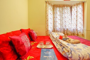 Quarto de hotel com colcha vermelha, cortinas florais e um tabuleiro com duas chávenas e um livro intitulado "Khana mein Kya hai?".