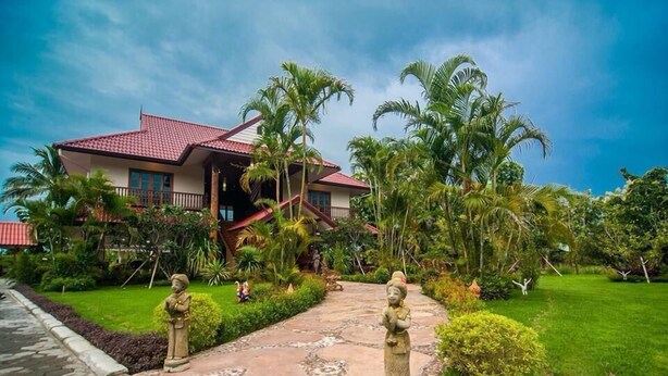 A house with a red roof, surrounded by lush greenery and palm trees. - Little Teak Home Stay (San Sai)