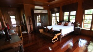 A spacious bedroom with a large bed, wooden flooring, and a window with a view of greenery. - Little Teak Home Stay (San Sai)