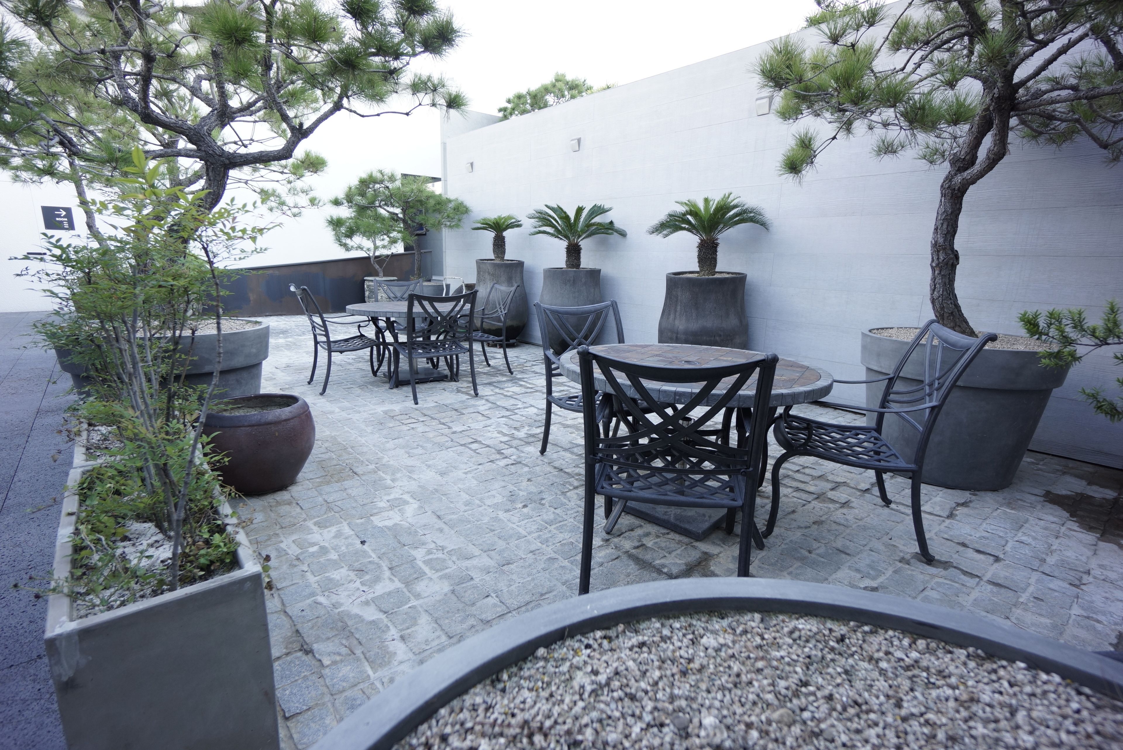 An outdoor patio area with stone flooring, multiple tables and chairs, and large planters with trees and shrubs.