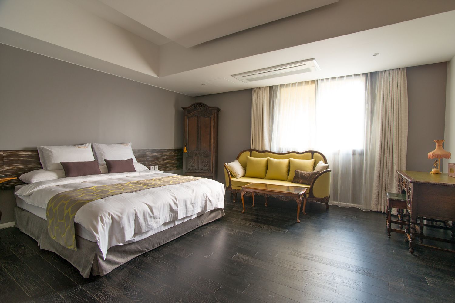 Un dormitorio con una cama grande, un armario de madera, un sofá amarillo, un escritorio de madera y un ventanal con cortinas.