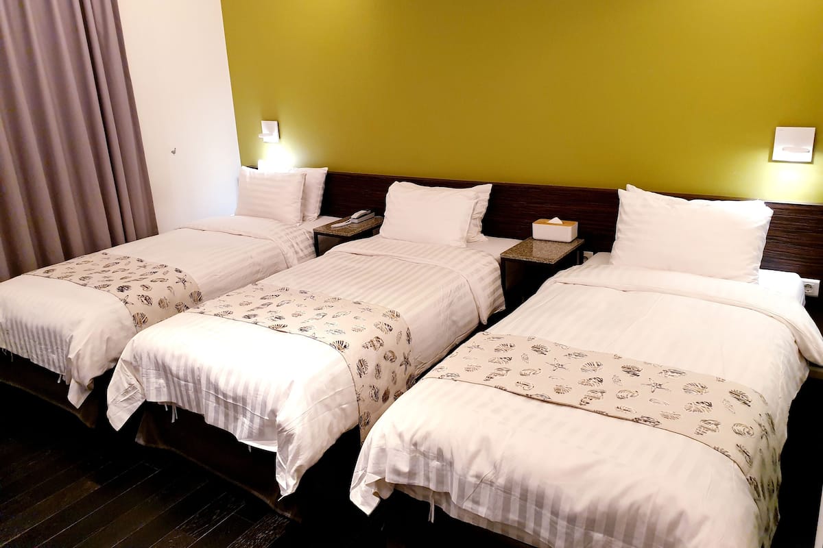Une chambre d’hôtel avec trois lits simples, chacun agrémenté d’une couvre-lit à motifs et de coussins blancs. Une table de chevet, sur laquelle se trouvent un téléphone et une boîte de mouchoirs, est placée d’un côté. La chambre est dotée d’une tête de lit en bois et d’un mur vert.