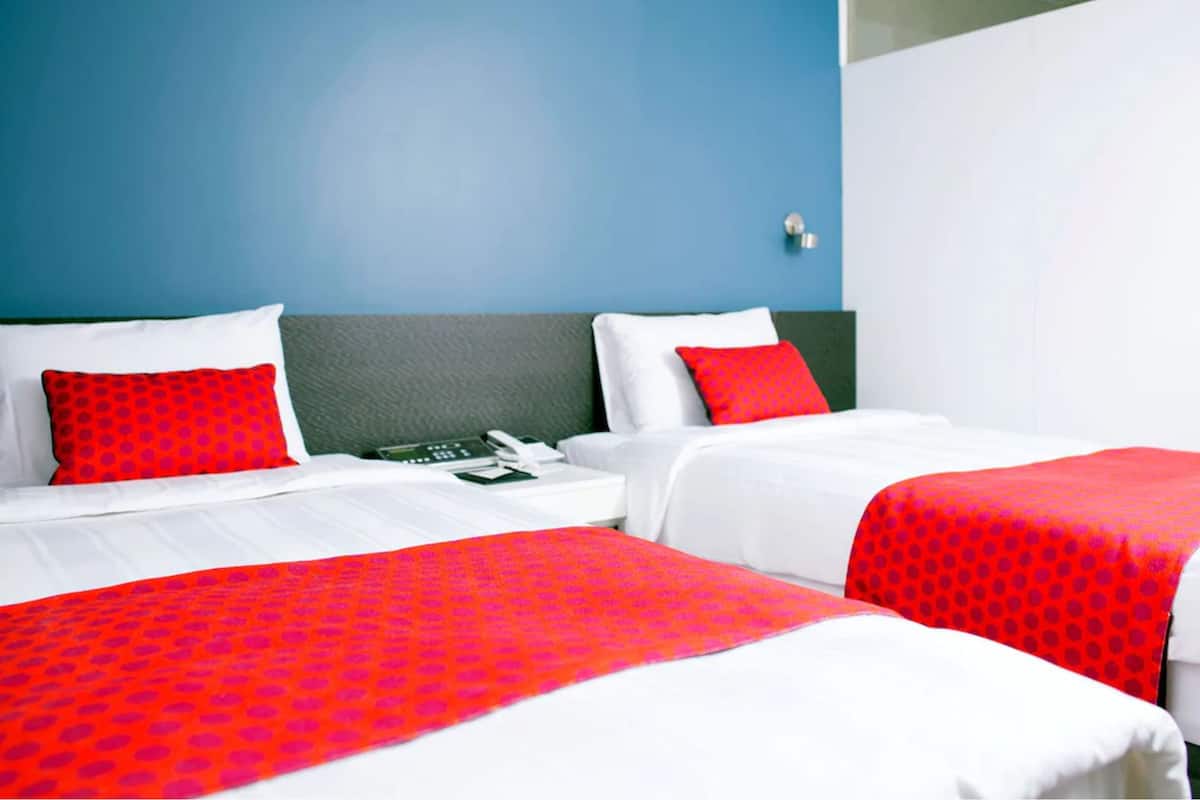 Une chambre d’hôtel avec deux lits, chacun agrémenté de draps blancs et de touches de couleur rouge, d’une tête de lit verte et d’un mur bleu.