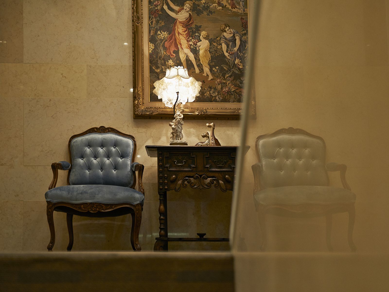 A room with two upholstered chairs, a wooden side table with a lamp, and a framed painting on the wall.