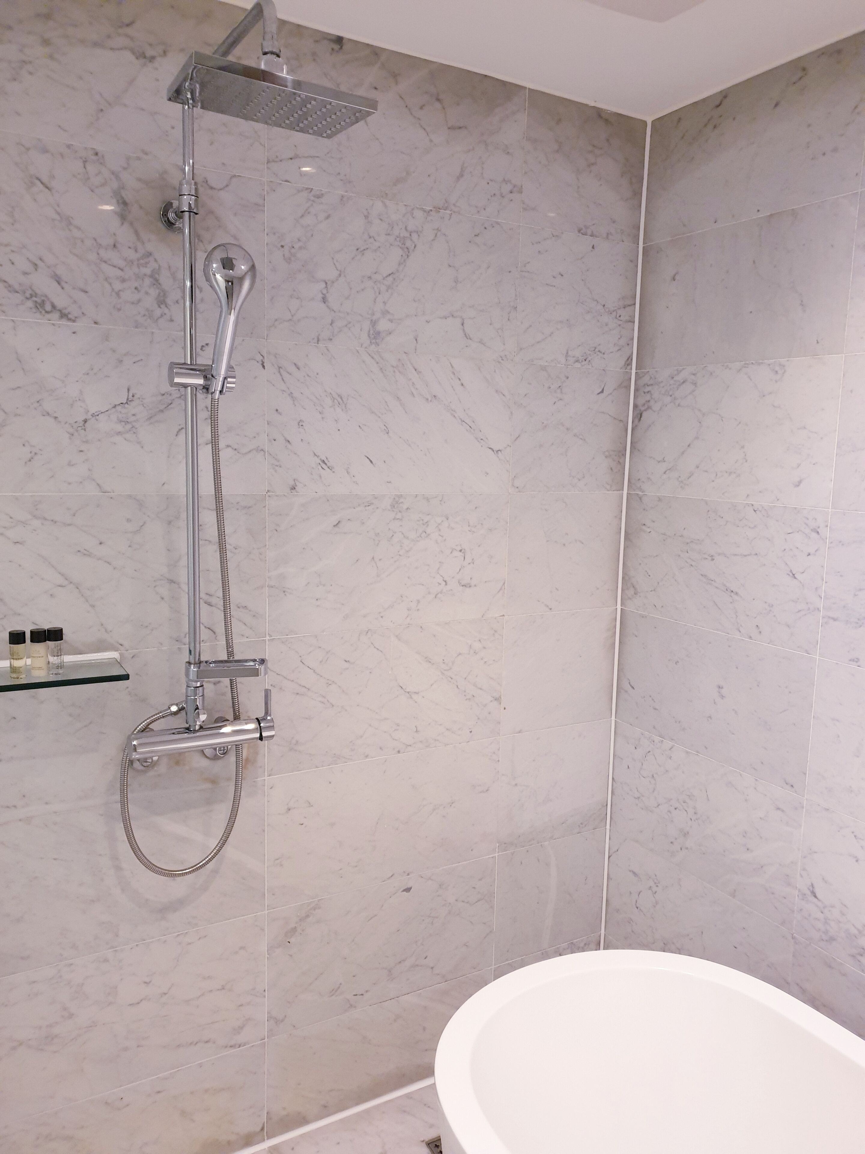 A modern bathroom with a shower, a glass shelf with toiletries, and a white bathtub.
