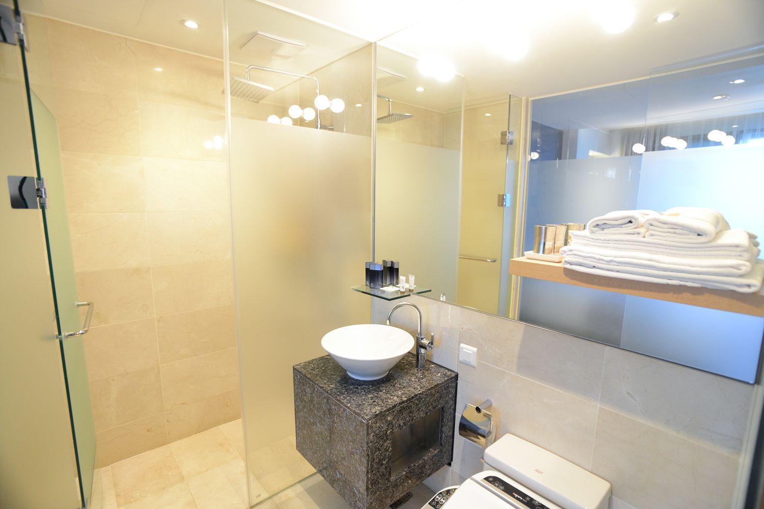 Un baño moderno con cabina de ducha de vidrio, encimera de granito y lavamanos blanco, y un perchero con toallas dobladas.
