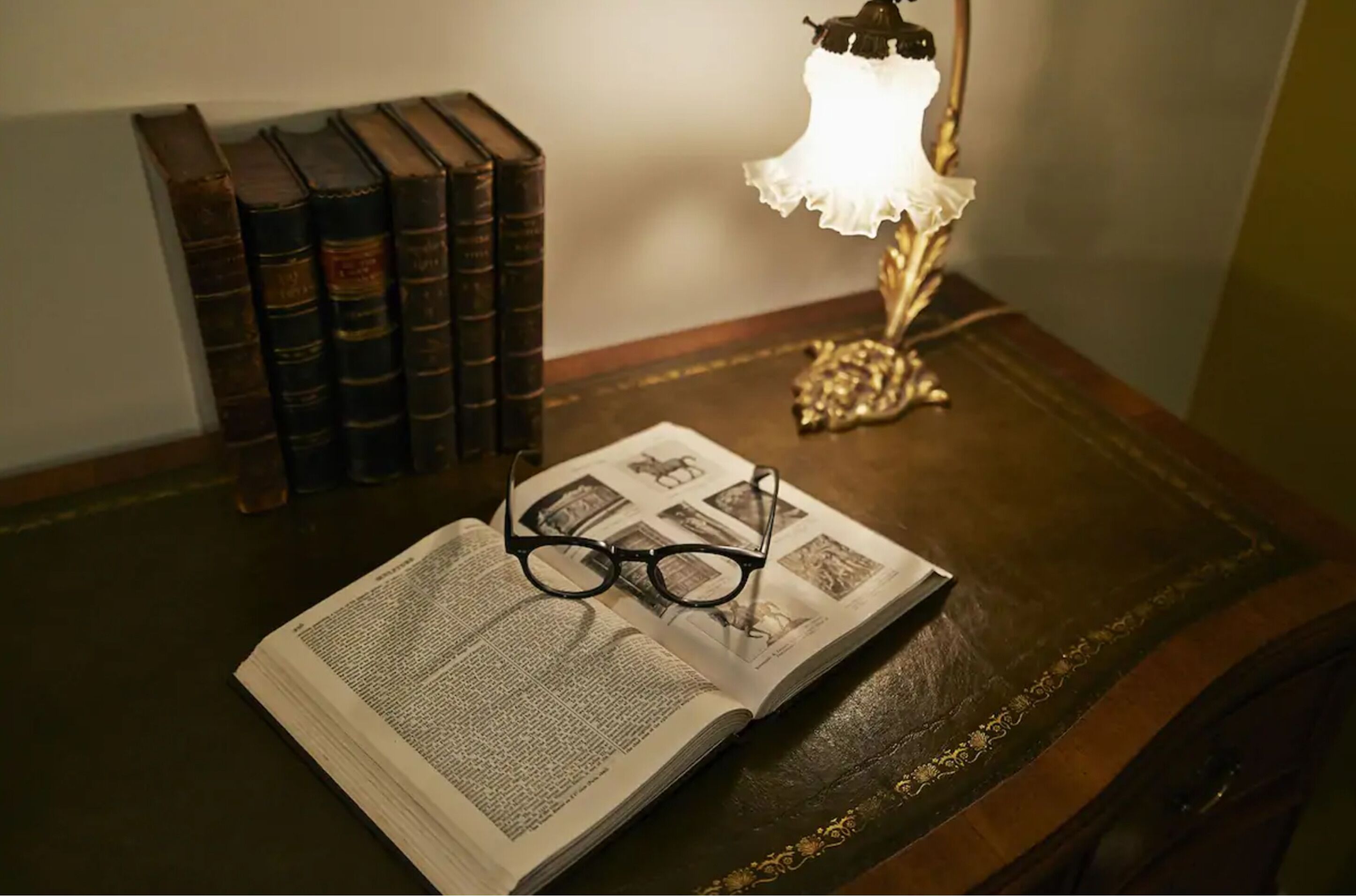 An open book with glasses on top, a lamp, and several books on a wooden table.