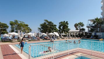 A pool area with multiple lounge chairs and umbrellas, surrounded by palm trees and a clear sky.