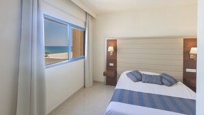 Una habitación de hotel con cama, mesita de noche, ventana con vista a la playa y lámpara sobre la mesita.