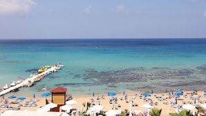 Una playa con filas de sillas de playa y sombrillas, un muelle con barcos y agua cristalina azul.