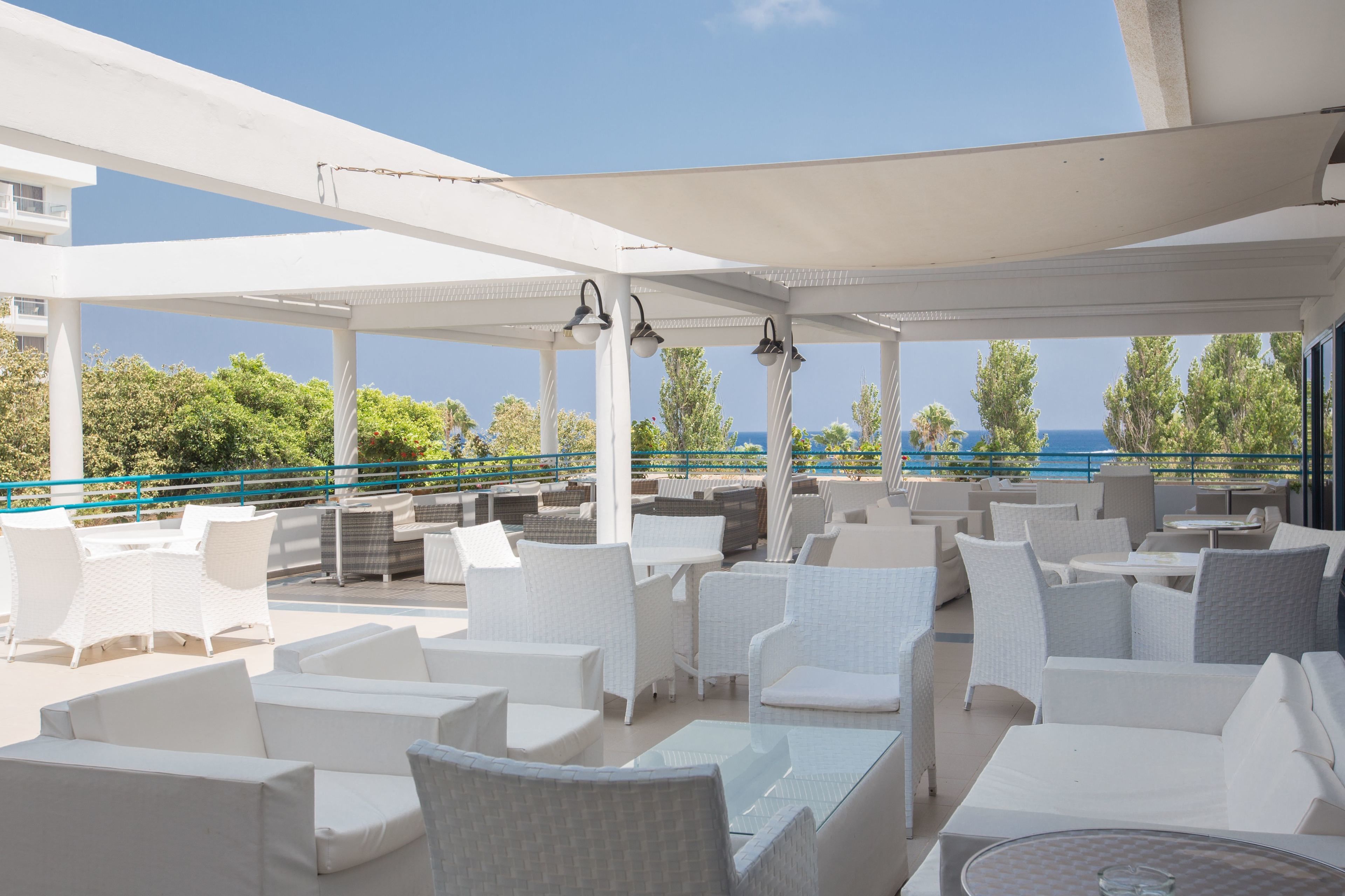 A covered outdoor seating area with white wicker furniture and tables, overlooking a body of water and greenery.