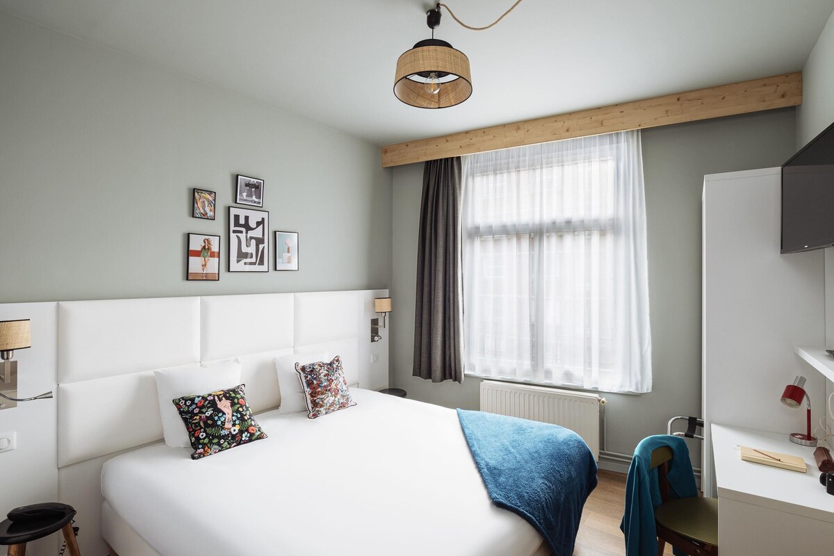 Un dormitorio con cama, mesita de noche, lámpara y ventana con cortinas.