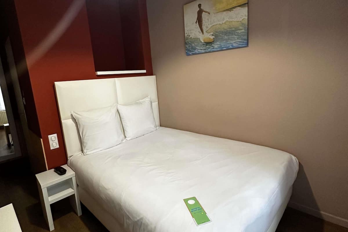 Una cama bien hecha con sábanas y almohadones blancos, una mesita de noche con un teléfono y un cuadro de un surfista en la pared.