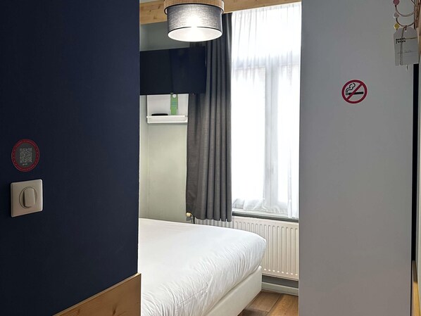 A bedroom with a bed, a window with curtains, a wall-mounted light fixture, a radiator, and a no-smoking sign.