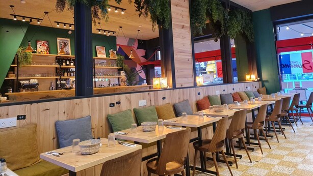 Un restaurante con largas mesas de madera, asientos con cojines verdes y plantas colgantes.