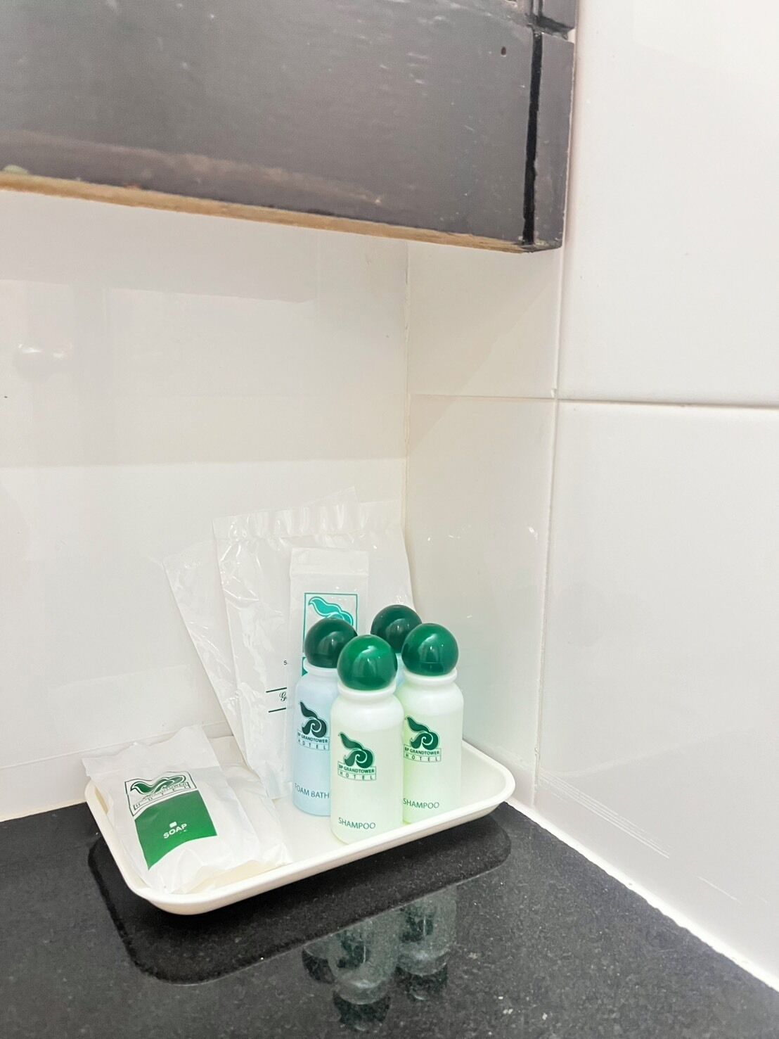 A bathroom with white tiles, a black countertop, and a tray holding four bottles of shampoo and a packet of soap.