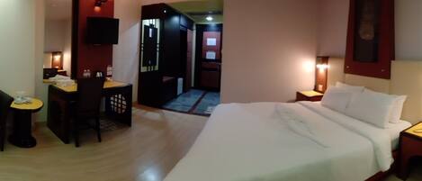 Una habitación de hotel con cama, mesitas de noche, un escritorio con silla, televisor y una puerta que da a un pasillo.