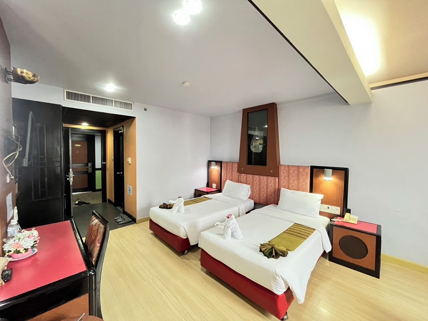 A hotel room with two beds, a desk, and a chair. There is a mirror above the beds and a small table with a red tablecloth.