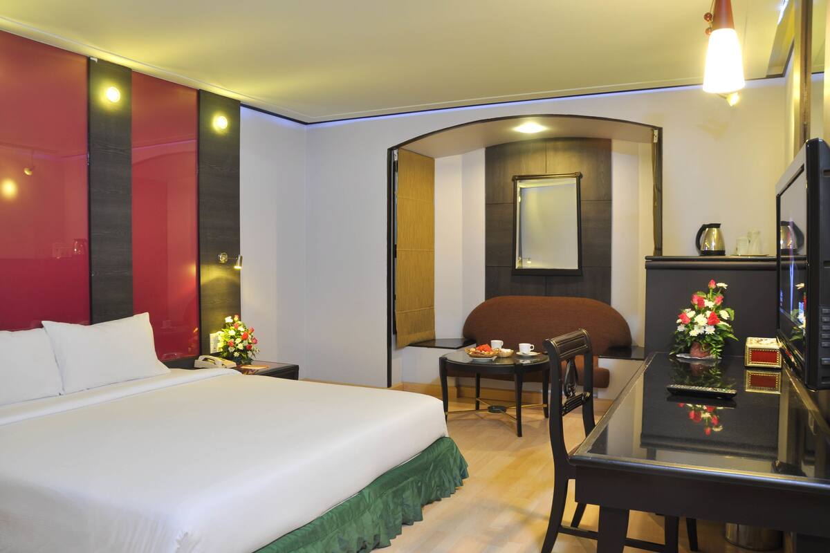 A hotel room with a large bed, a desk with a chair, a small table with a cup and flowers, and a TV on a stand.